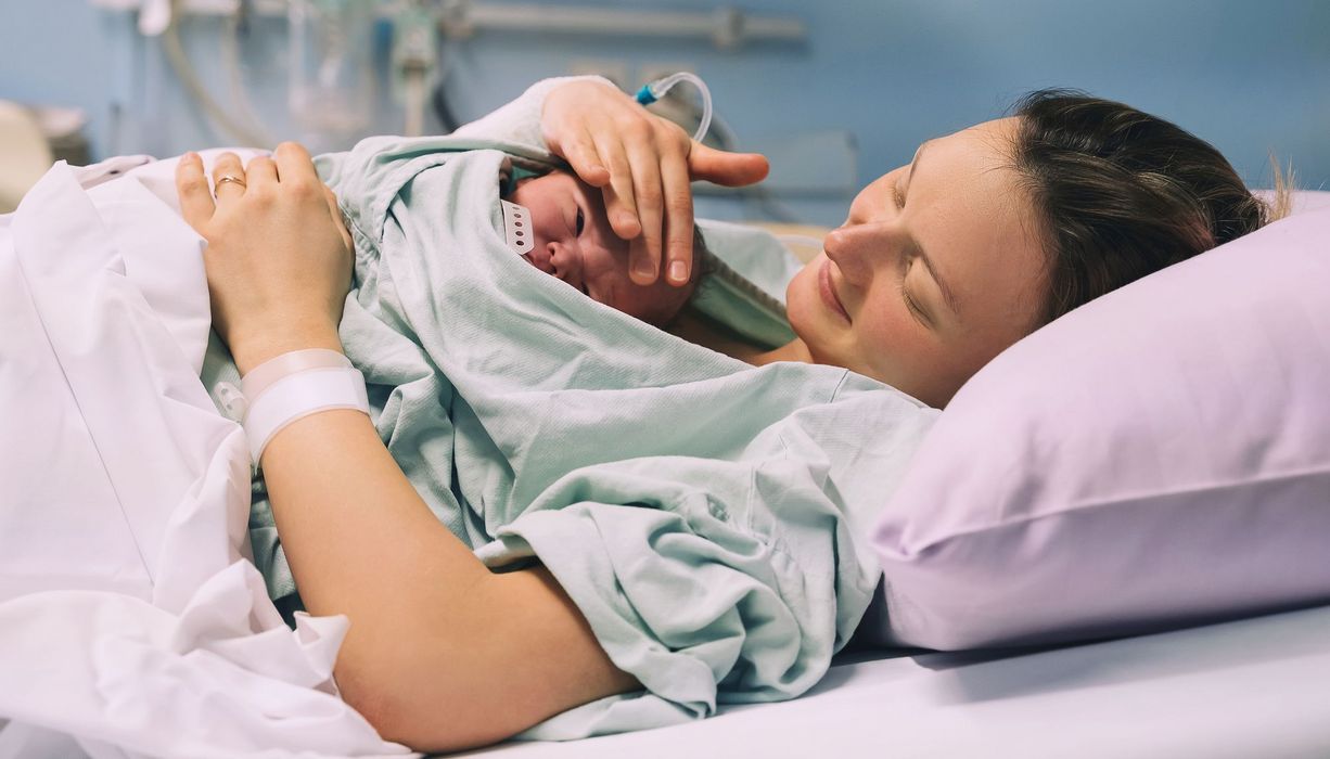 Frau liegt nach der Geburt mit ihrem Neugeborenen im Krankenhaus im BNett. Sie streichelt lächelnd die Stirn des Babys.