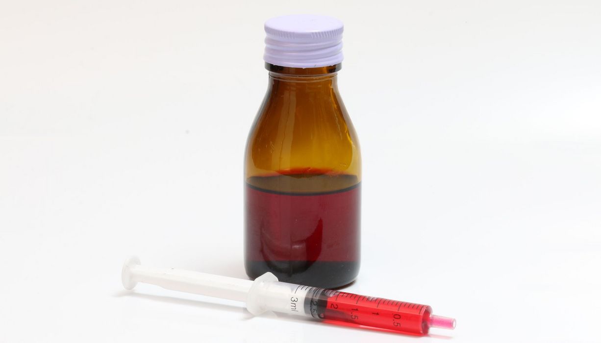 Eine, mit roter Flüssigkeit gefüllte, Pipette liegt vor weißem Hintergrund neben einer Braunglasflasche. 