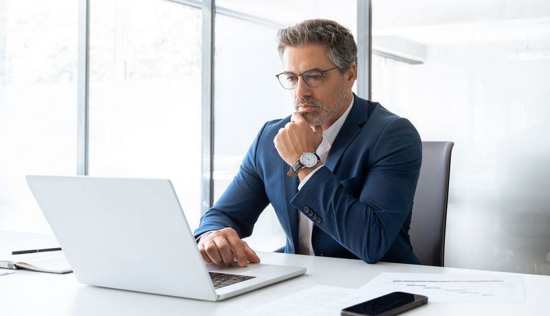Nachdenklicher Mann in Anzug zwischen 45 und 50 am Laptop im Büro.