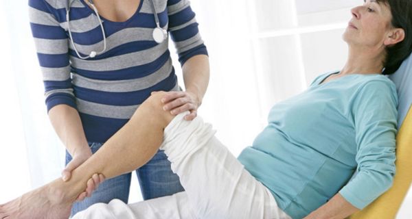 Frau mit Knieschmerzen (um die 50) auf Arztliege bekommt von kopfloser Ärztin (Stethoskop um den Hals) linkes Bein bewegt, eine Hand am Knie