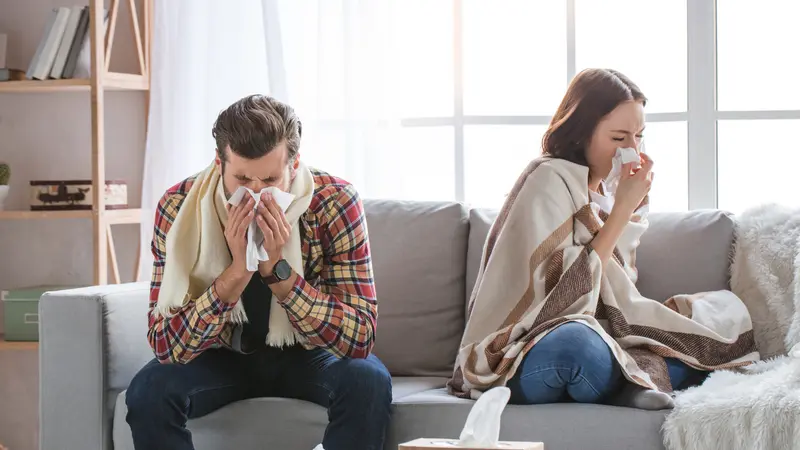 Mann und Frau sitzen – eingehüllt in Decke und Schal – auf dem Sofa, niesen und putzen sich die Nase.