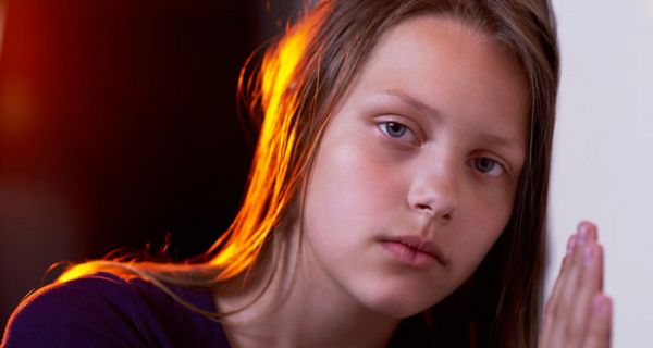 Sehr traurig blickendes Mädchen, ca. 11 Jahre alt, mit langen braunen Haaren vor dunklem Hintergrund, eine Hand und Wange an eine helle Wand gelehnt