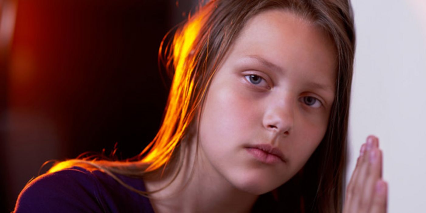Sehr traurig blickendes Mädchen, ca. 11 Jahre alt, mit langen braunen Haaren vor dunklem Hintergrund, eine Hand und Wange an eine helle Wand gelehnt