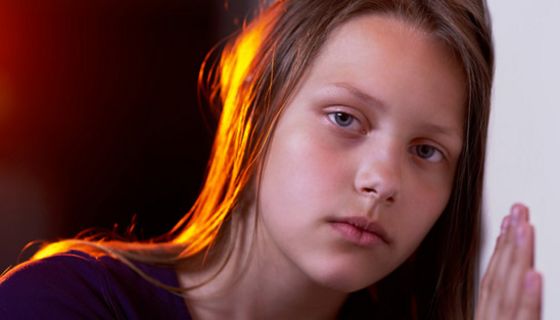 Sehr traurig blickendes Mädchen, ca. 11 Jahre alt, mit langen braunen Haaren vor dunklem Hintergrund, eine Hand und Wange an eine helle Wand gelehnt