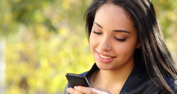 Außenaufnahme, Portrait bis Schulterpartie: Junge attraktive Frau, lange, dunkle, glatte Haare, schwarze Lederjacke, schaut lächelnd auf ein Smartphone