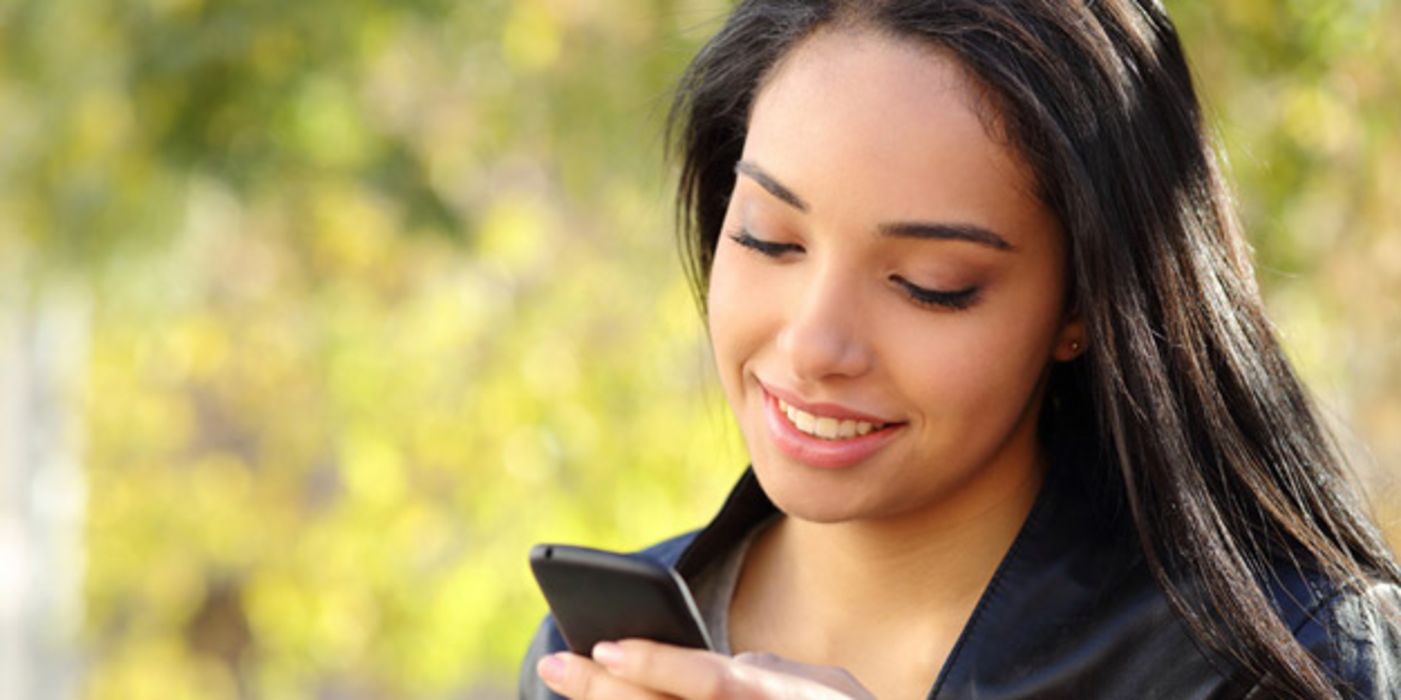 Außenaufnahme, Portrait bis Schulterpartie: Junge attraktive Frau, lange, dunkle, glatte Haare, schwarze Lederjacke, schaut lächelnd auf ein Smartphone