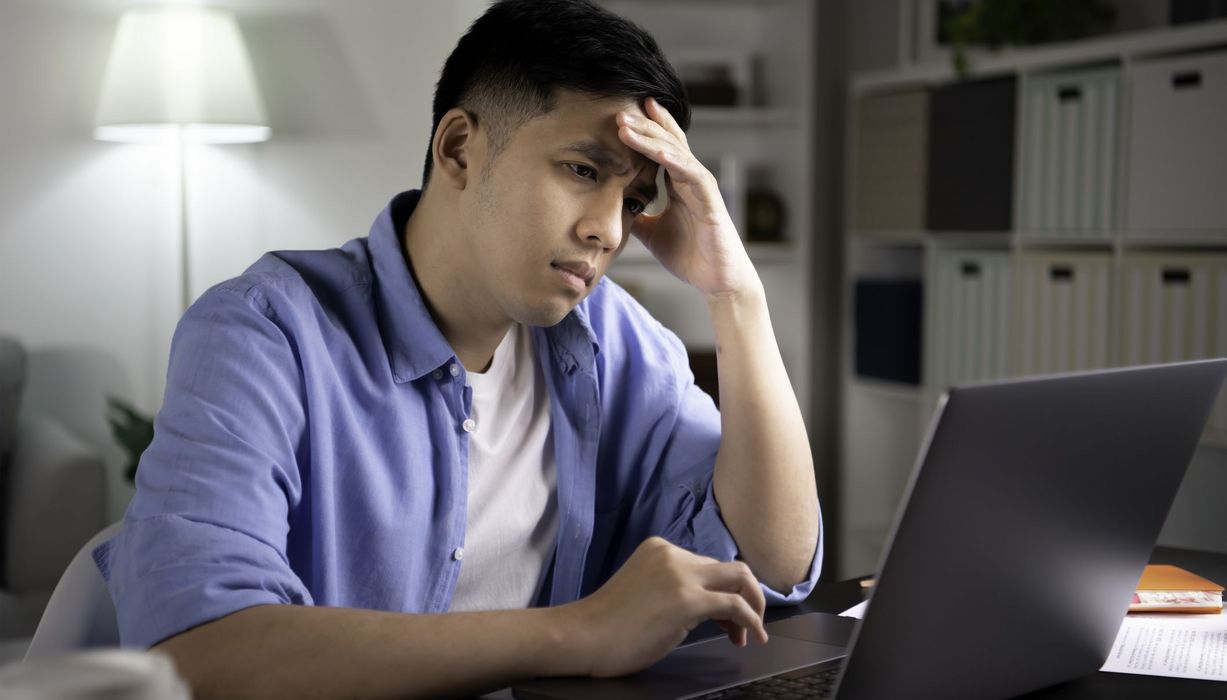 Junger Mann mit Augenringen hält sich die Stirn und blickt betroffen auf einen Laptopbildschirm.