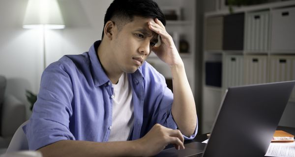 Junger Mann mit Augenringen hält sich die Stirn und blickt betroffen auf einen Laptopbildschirm.