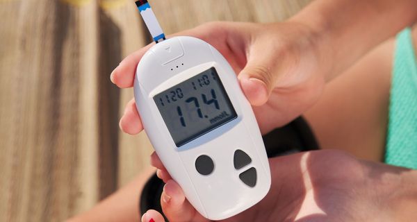 Blutzuckermessung am Strand: Eine Frau hält das Messgerät mit einem blutenden Finger im Bikini (nur Hände, Oberschenkel und Bikini-Hösschen sind zu sehen)