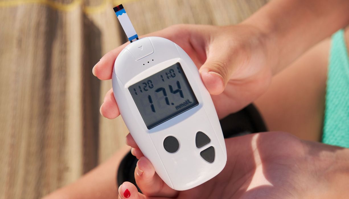 Blutzuckermessung am Strand: Eine Frau hält das Messgerät mit einem blutenden Finger im Bikini (nur Hände, Oberschenkel und Bikini-Hösschen sind zu sehen)