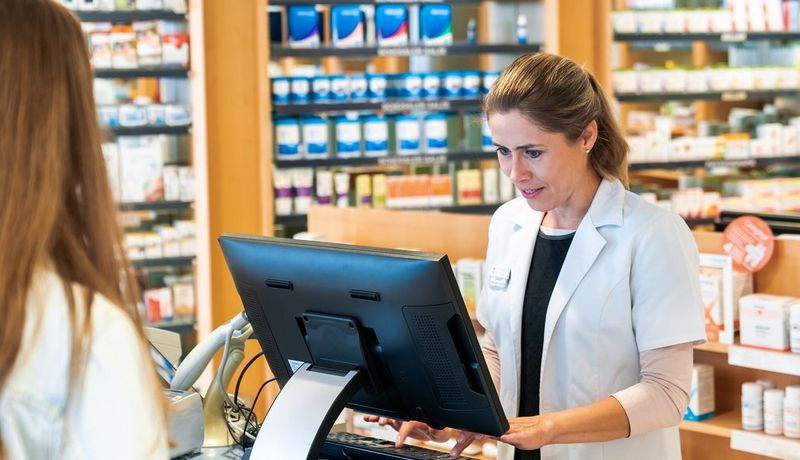 Apothekerin gibt etwas in einen Computer ein, von hinten angeschnitten sieht man eine Kundin.
