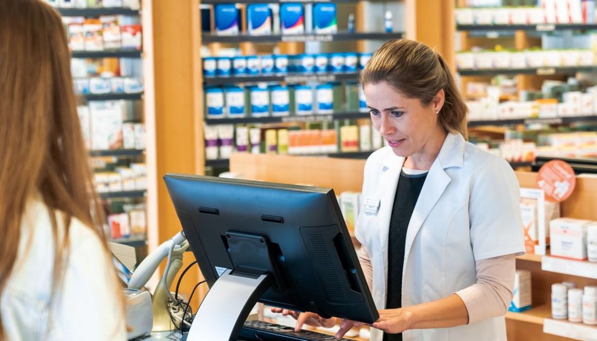 Apothekerin gibt etwas in einen Computer ein, von hinten angeschnitten sieht man eine Kundin.