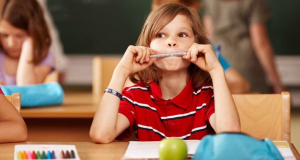 Kind, schulterlange Haare (Junge?), rotes Poloshirt mit weißen Streifen, am Stift kauend und nach oben schauend am Pult in der Schule sitzend, Tafel im Hintergrund