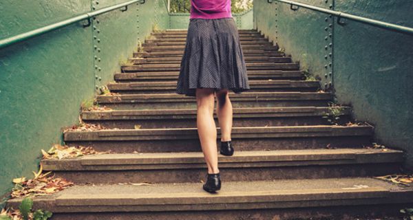 Rückansicht Frau im graugemusterten, knielangen Rock, zyklamfarbenes Oberteil z.T. zu sehen, läuft in einem Park eine Treppe hoch