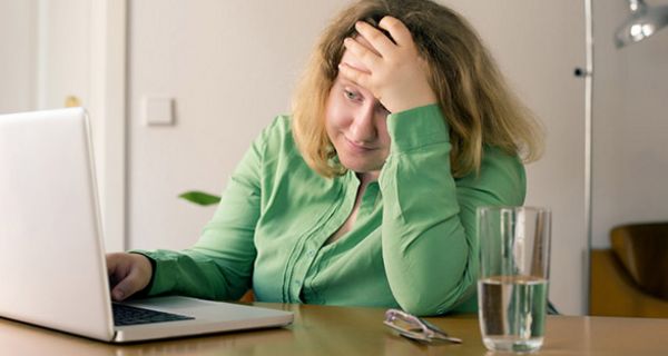 Übergewichtige Frau ca. Ende 20, lange Haare, grünes Sweatshirt, an Schreibtisch mit Laptop, gestresst schauend, Kopf auf eine Hand gestützt