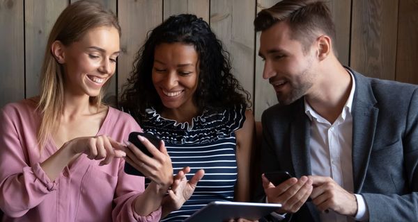 Zwei Frauen und ein Mann mit Smartphones bzw. Tablet in den Händen reden miteinander und schauen gemeinsam auf ein Smartphone. 