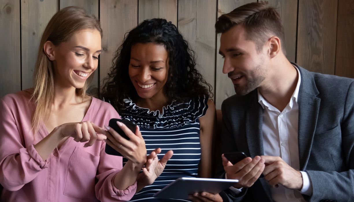 Zwei Frauen und ein Mann mit Smartphones bzw. Tablet in den Händen reden miteinander und schauen gemeinsam auf ein Smartphone. 