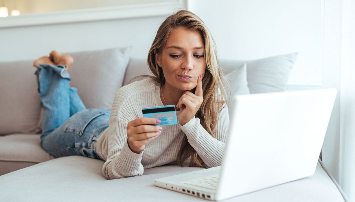 Junge Frau schaut zweifelnd auf ihre Kreditkarte, vor ihr steht ihr aufgeklappter Laptop.