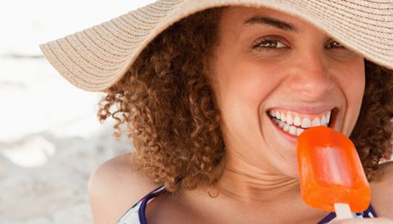 Nahaufnahme Gesicht dunkelhaarige Frau, großer Strohhut, beißt genüsslich in ein Fruchteis (orangefarben)