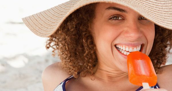 Nahaufnahme Gesicht dunkelhaarige Frau, großer Strohhut, beißt genüsslich in ein Fruchteis (orangefarben)