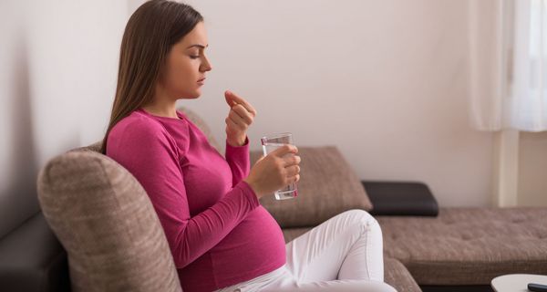 Schwangere Frau hält in der einen Hand ein Schmerzmittel und in der anderen Hand ein Glas Wasser.