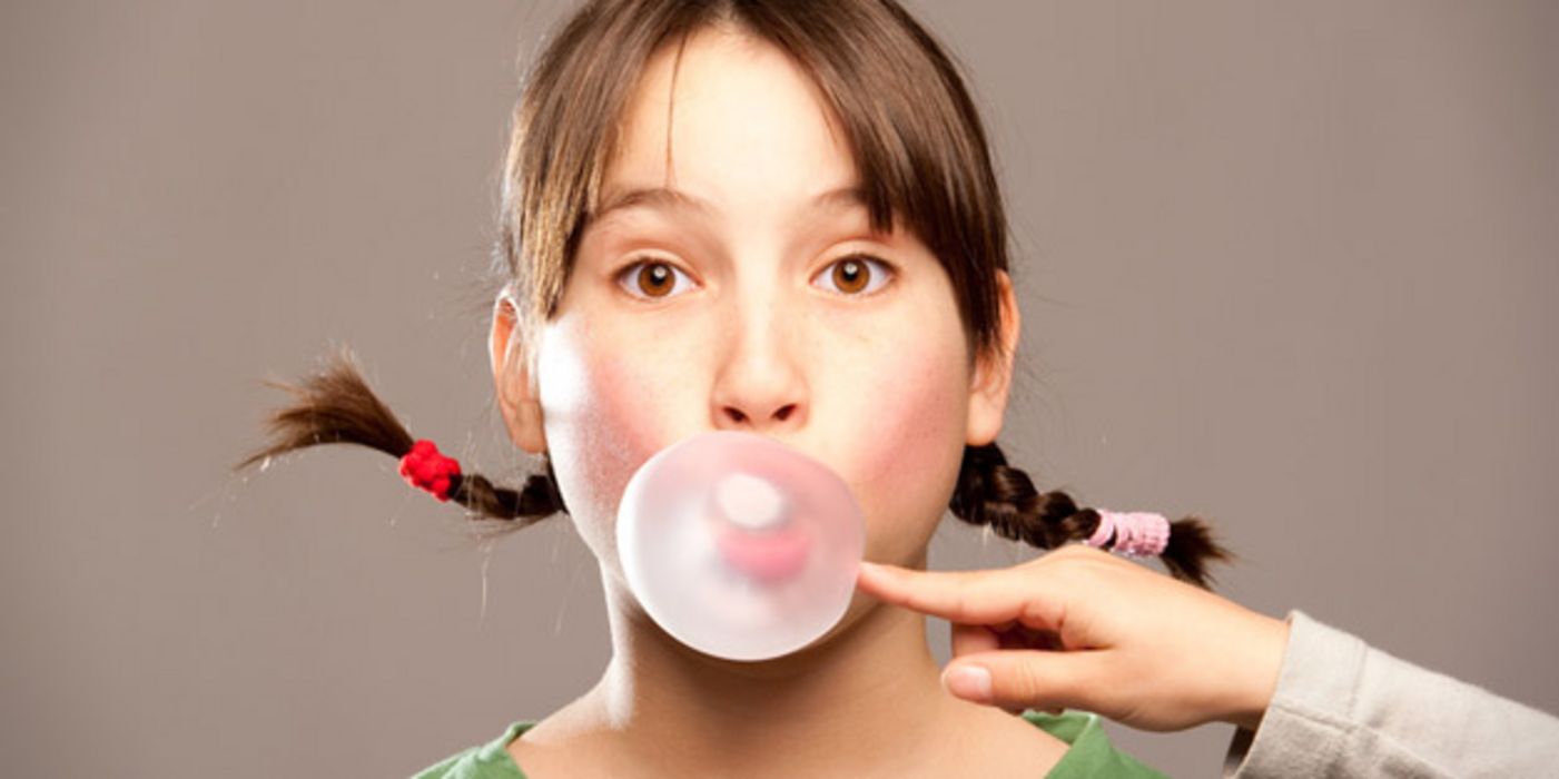 Mädchen mit Zöpfen macht Kaugummiblase, Hand aus dem Off versucht, diese zum Platzen zu bringen