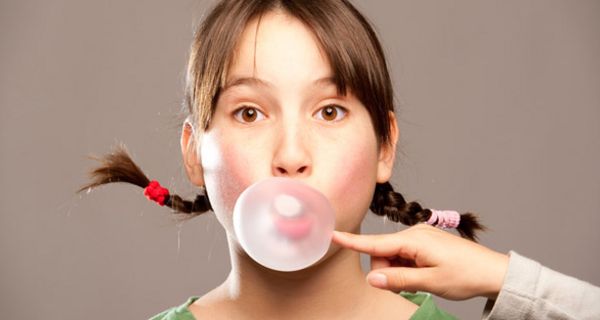 Mädchen mit Zöpfen macht Kaugummiblase, Hand aus dem Off versucht, diese zum Platzen zu bringen