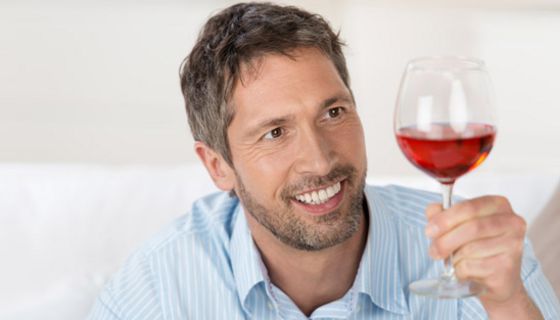 Mann mit Dreitagebart im hellblau-weiß gestreiften Hemd betrachtet lächelnd ein bauchiges Glas, gefüllt mit Rotwein