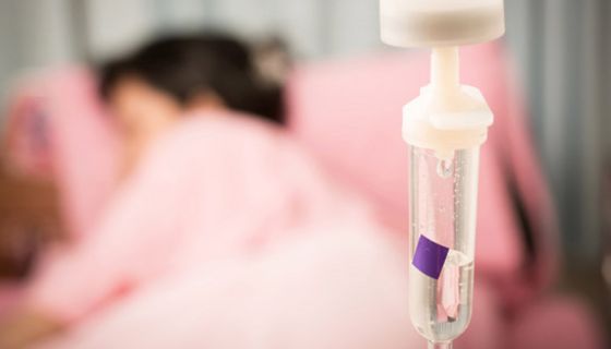 Krankenhaus; Infusionstropf im Vordergrund, im Hintergrund liegt schemenhaft Mensch im Krankenbett