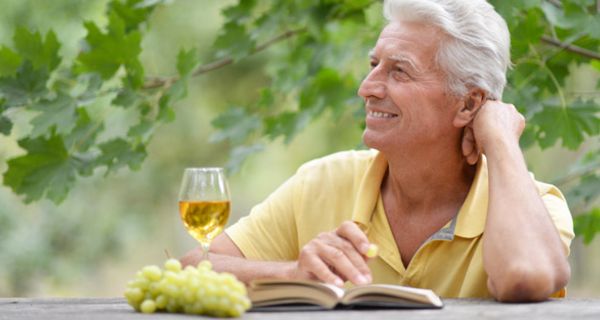 Porträtfoto weißhaariger Mann, gelbes Poloshirt, linker Elenbogen auf Tisch gestützt, Hand am Gesicht, Weißwein und Buch vor sich, Laubbaum hinter sich, blickt leicht nach oben ins Off