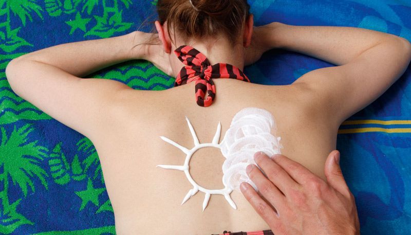Frau im Bikini liegt auf dem Bauch und wird mit Sonnencreme eingecremt