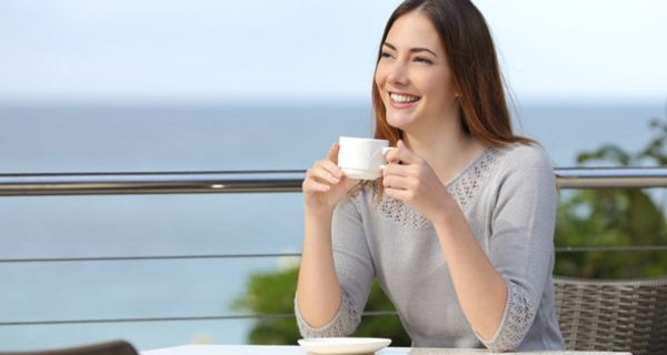 Junge Frau in den 20ern, leichter hellgrauer Sommerpulli, lange, braune Haare, lachend auf einer Sonnenterrasse am Meer, eine Tasse Kaffee in den Händen