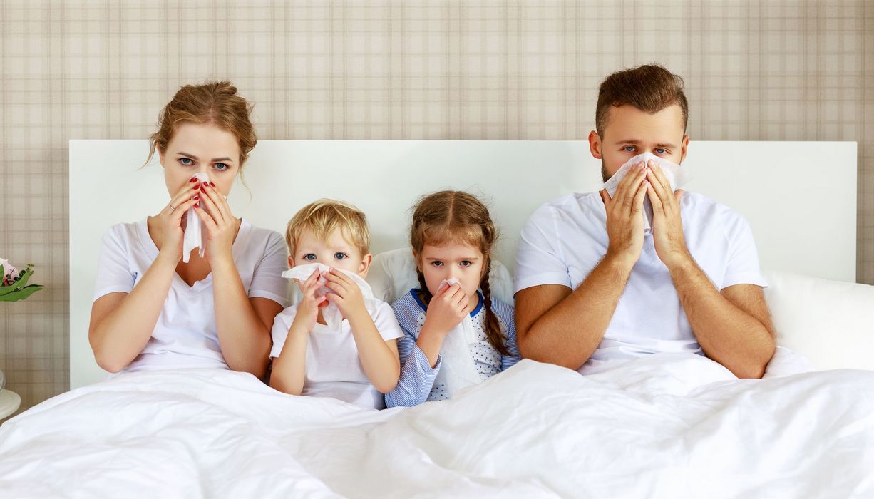 Mutter, zwei Kinder und Vater, liegen alle zusammen im Bett und putzen sich die Nase.