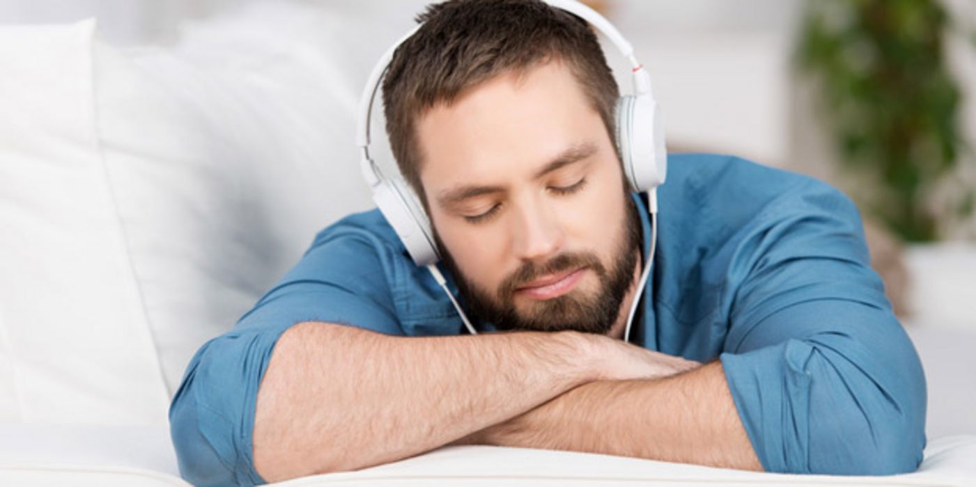 Junger, bärtiger Mann mit geschlossenen Augen liegt auf einer Couch und hört Musik über Kopfhörer