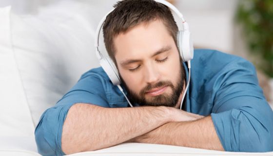 Junger, bärtiger Mann mit geschlossenen Augen liegt auf einer Couch und hört Musik über Kopfhörer