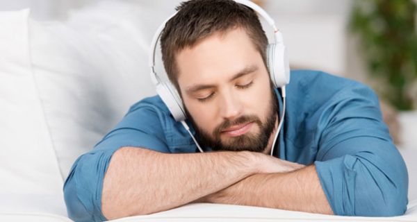 Junger, bärtiger Mann mit geschlossenen Augen liegt auf einer Couch und hört Musik über Kopfhörer