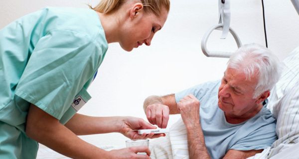 Senior im Krankenbett nimmt eine Tablette aus einer Dosette, die ihm eine junge Krankenschwester hinhält