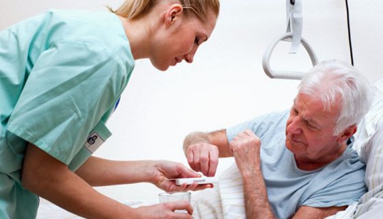 Senior im Krankenbett nimmt eine Tablette aus einer Dosette, die ihm eine junge Krankenschwester hinhält