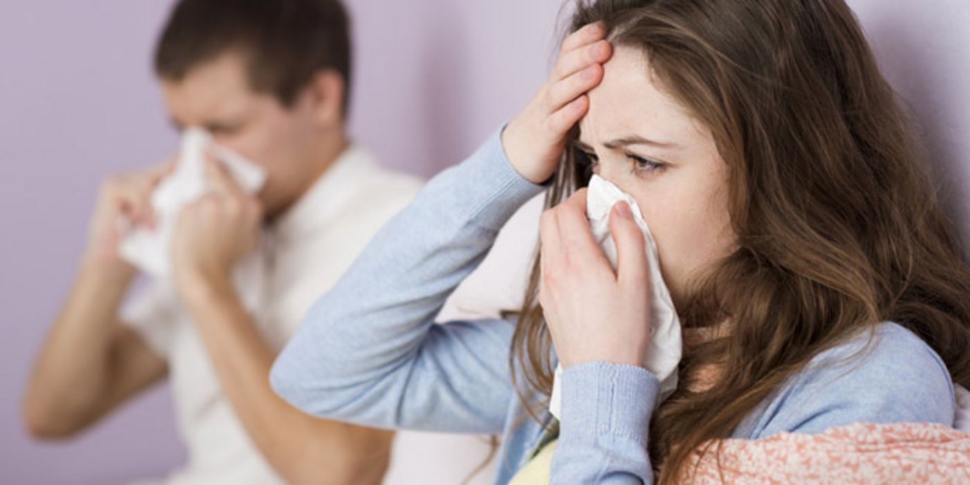 Grippe, Frau und Mann putzen sich die Nasen, Frau im Vordergrund hält sich Hand an die Stirn 