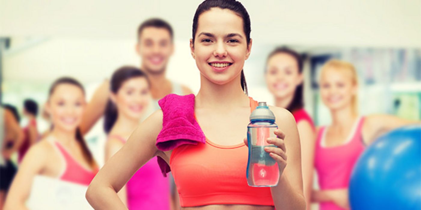 Fitnessraum: Vorne Teenager (weibl.), bauchfreies Top, graue Sporthose am Ansatz zu sehen, Wasserflasche in einer Hand; im Hintergrund in Unschärfe mehrere junge Leute