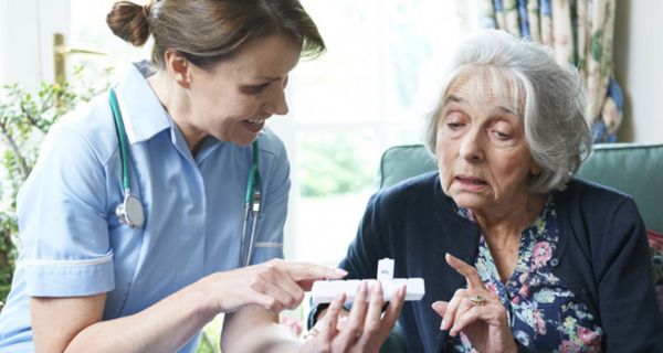 Seniorin bekommt von Pflegerin die Tabletteneinnahme mit einer Dosette erklärt