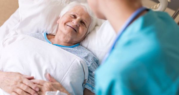 Lächelnde ältere Frau im Krankenhausbett, sie bekommt von einer Ärztin die Hand gehalten