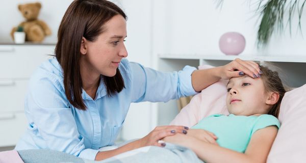 Kleines Mädchen liegt im Bett, eine junge Frau schaut sie besorgt an, legt ihr die Hand an die Stirn. 