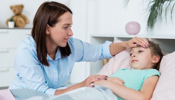 Kleines Mädchen liegt im Bett, eine junge Frau schaut sie besorgt an, legt ihr die Hand an die Stirn. 