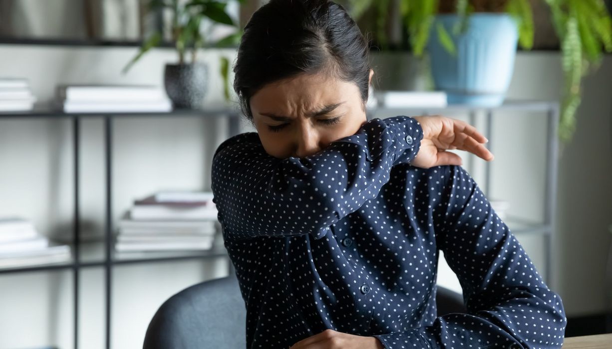 Frau, hustet in ihren Ellenbogen und sitzt im Büro.