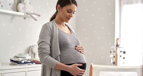 Eine hochschwangere Frau streichelt sich im Babyszimmer den Bauch und lächelt.