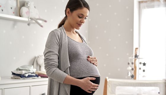 Eine hochschwangere Frau streichelt sich im Babyszimmer den Bauch und lächelt.