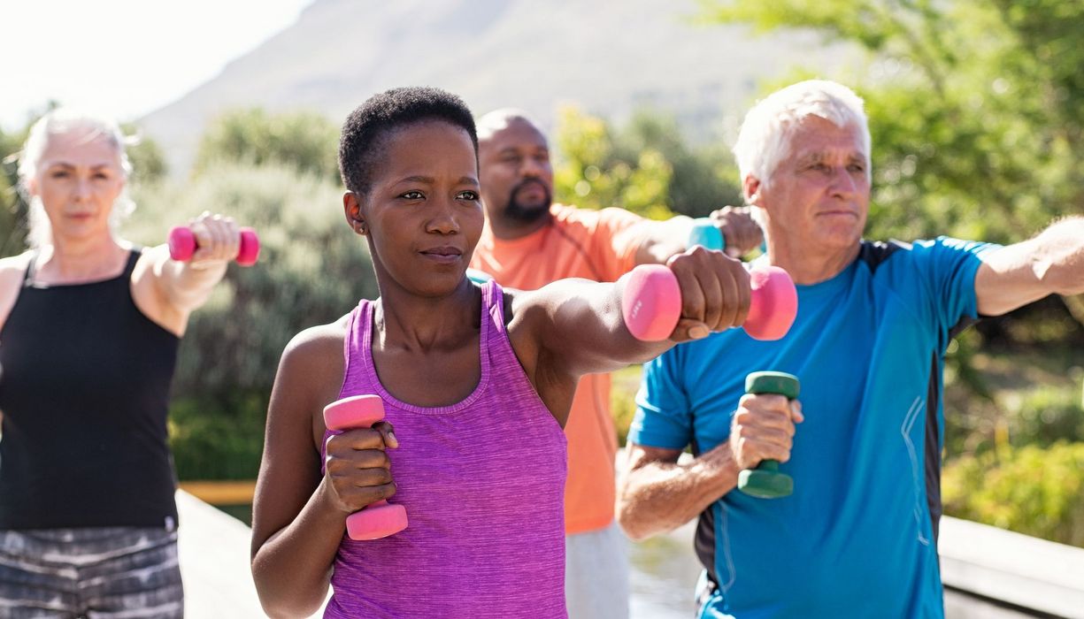 Vier Erwachsene unterschiedlichen Alters, zwei Frauen, zwei Männer, trainieren unter freiem Himmel mit Hanteln.