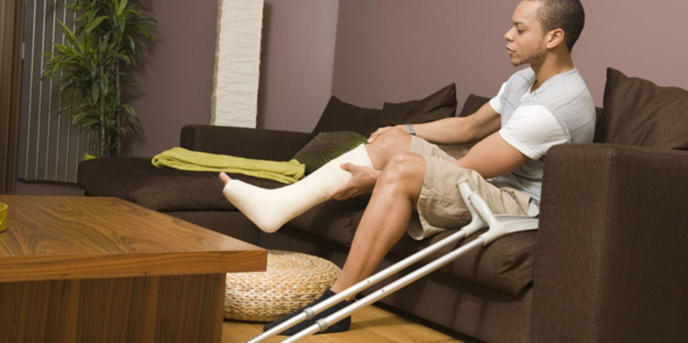 Junger Mann sitzt auf einem Sofa mit Gipsbein und Krücken neben sich.