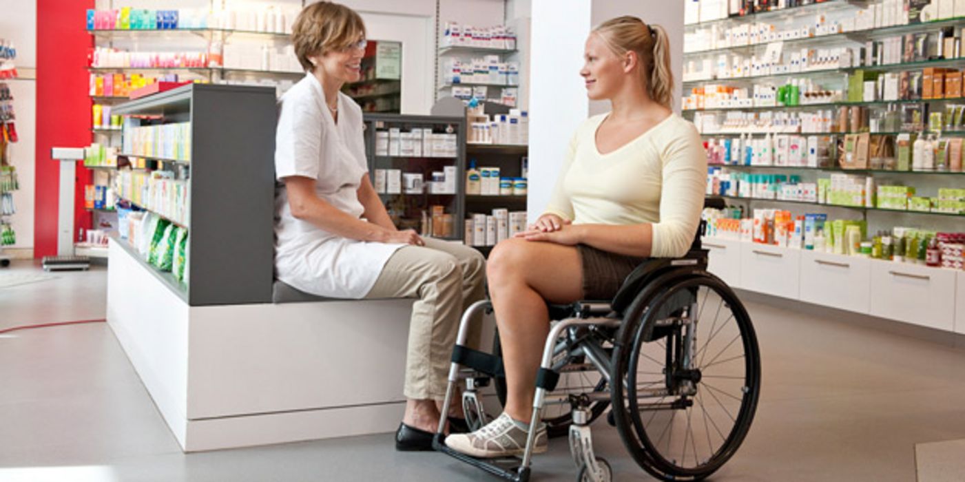 Frau im Rollstuhl und Apothekerin im Gespräch in einer Apotheke